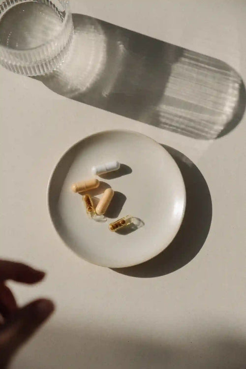 Assiette blanche contenant plusieurs gélules de compléments alimentaires Jolly Mama posée sur une table ensoleillée, à côté d’un verre d’eau.