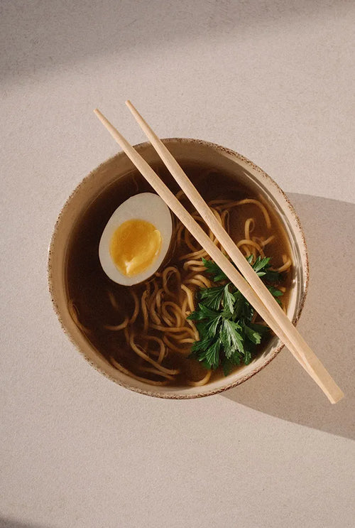 Bol de bouillon fumant aux nouilles, œuf dur et herbes fraîches, avec baguettes posées – préparation saine et réconfortante à base de Broth, idéale pour la récupération post-partum et l’hydratation.