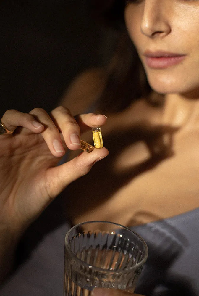 Femme tenant délicatement une capsule dorée de complément alimentaire avec un verre d’eau – illustration d’un supplément naturel pour la fertilité féminine et le soutien hormonal.