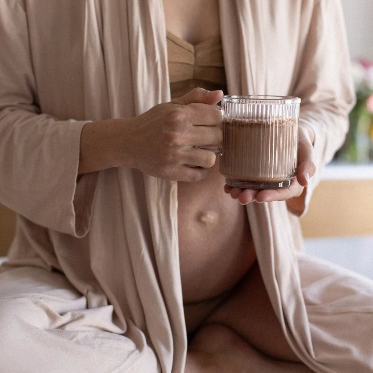 Femme enceinte assise en peignoir beige, tenant une tasse de boisson chaude préparée avec Mamaload, la poudre cacao-collagène de Jolly Mama. Un rituel bien-être qui soutient la peau, les cheveux et les tissus pendant et après la grossesse.

