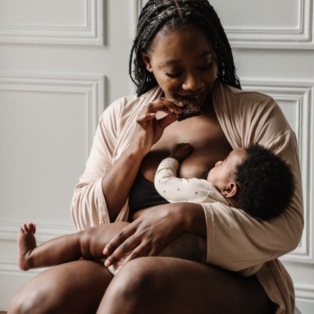 Femme en train d’allaiter son bébé, mangeant un encas Jolly Mama adapté à l’allaitement